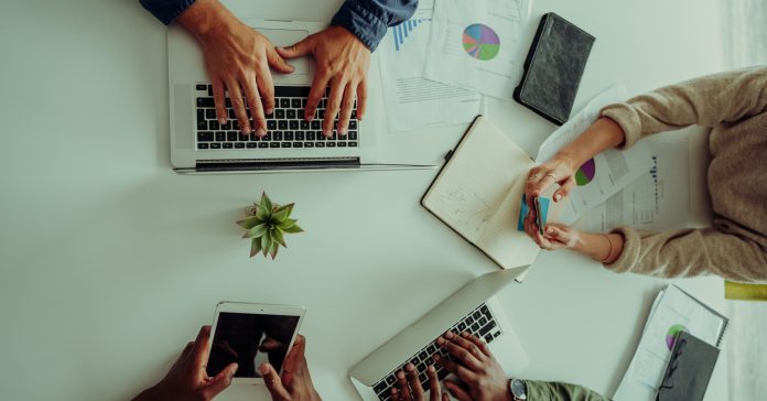 Group of mixed race workers gather around table having discussing high angle shot in conference room working off laptops and searching through documents. High quality photo